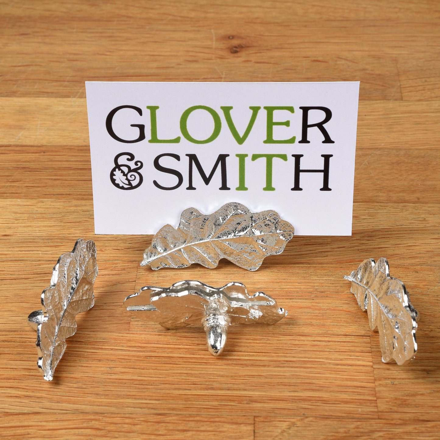 A set of four silver-colored pewter oak leaf place card holders displayed on a wooden surface with a business card branded 'Glover & Smith'.
