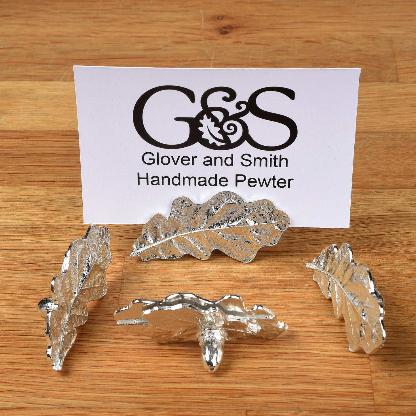 A set of four silver-colored pewter oak leaf place card holders displayed on a wooden surface with a business card branded 'Glover & Smith'.