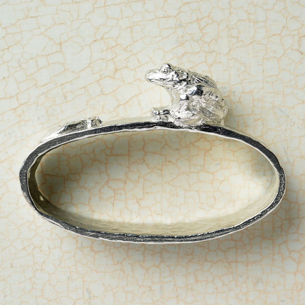 An oval-shaped pewter napkin ring featuring a textured design with a frog chasing an insect on top.