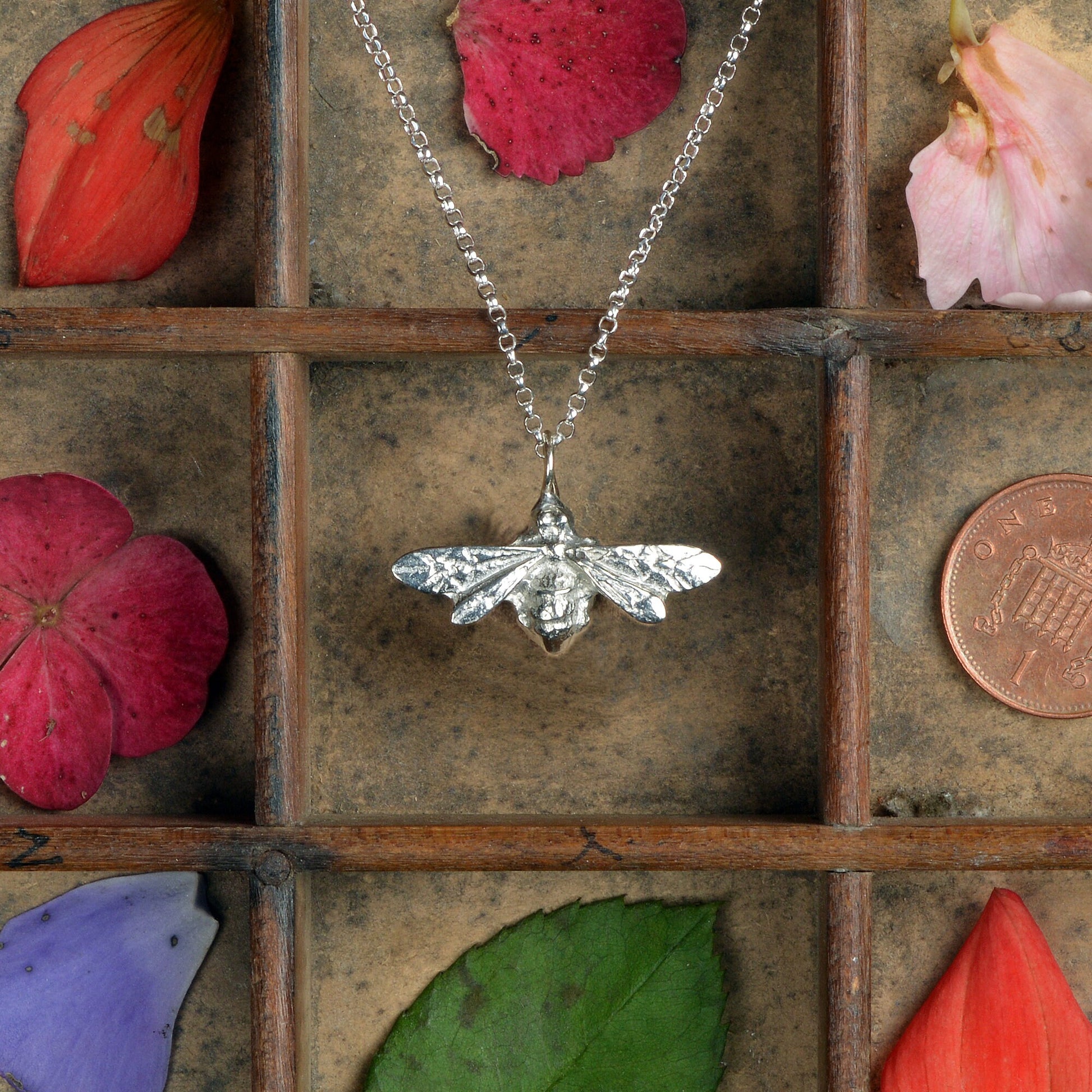 A silver-colored pewter honey bee pendant necklace with delicately detailed wings, displayed on a rustic wooden background.