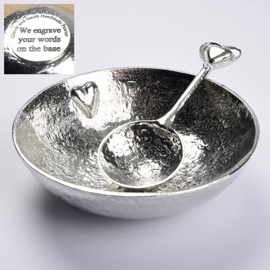 A textured pewter bowl with a raised small heart on the inner surface. It comes with a spoon with 2 hearts on the finial. One heart is within the other providing a tactile handle. The bowl of the spoon is round echoing the shape of the bowl. We engrave the base with your words.