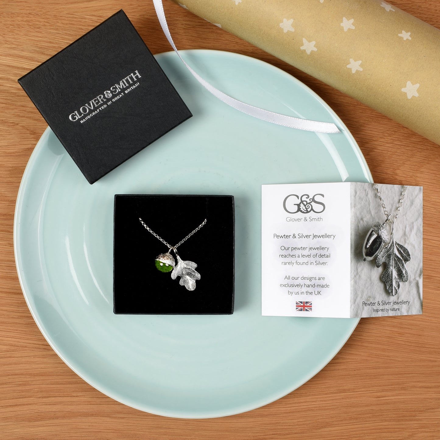 A pewter leaf pendant necklace with a green peridot accent is displayed in a black gift box on a light blue plate, alongside a black Glover & Smith box, a branded card, and a rolled gold gift wrap on a wooden surface.