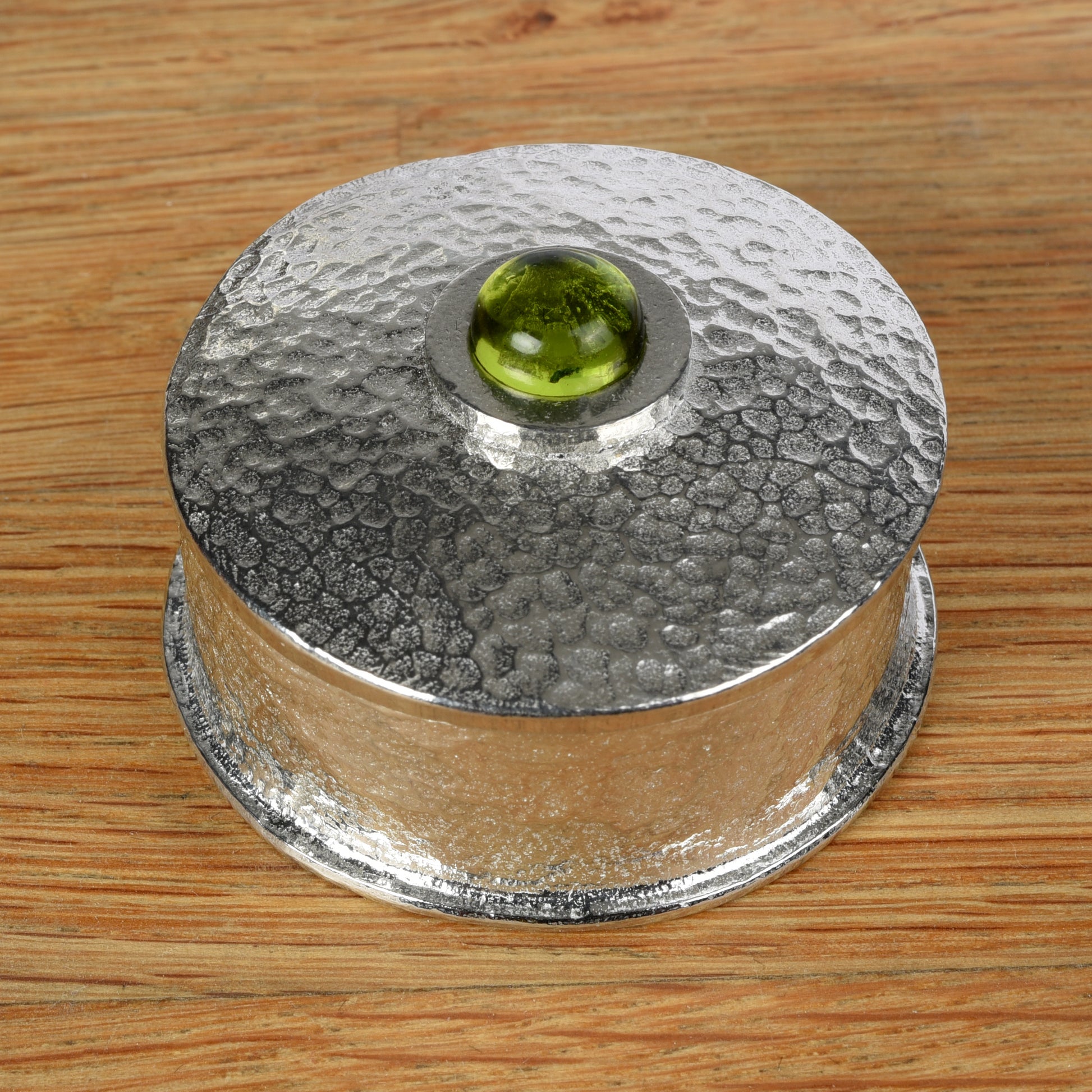 A hammered silver-toned pewter box set on a wooden surface. The lid features a circular cutout in the center revealing a peridot stone inlay. August birthstone gifts