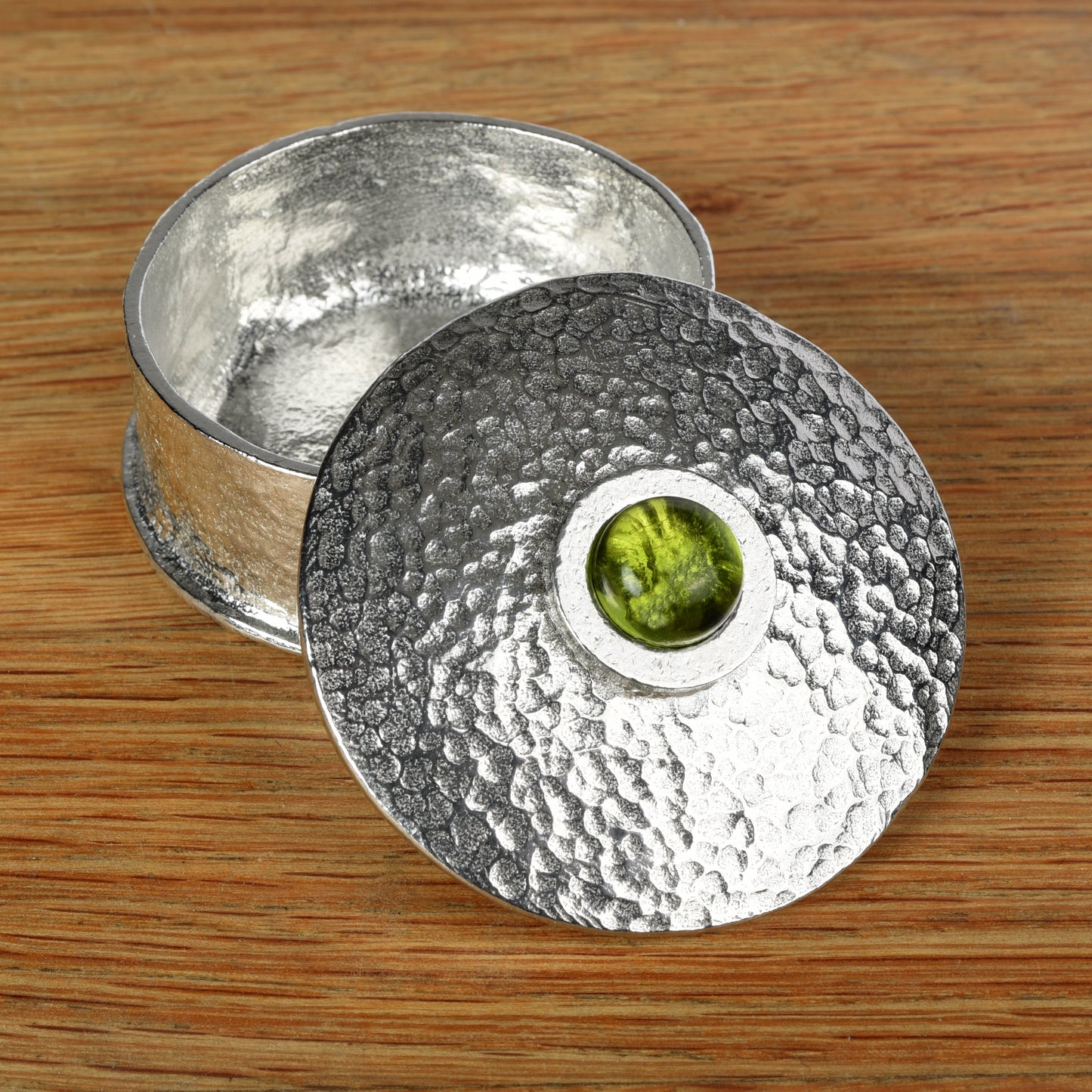 A hammered silver-toned pewter box with its lid leaning against the base, set on a wooden surface. The lid features a circular cutout in the center revealing a peridot stone inlay. August birthstone gifts