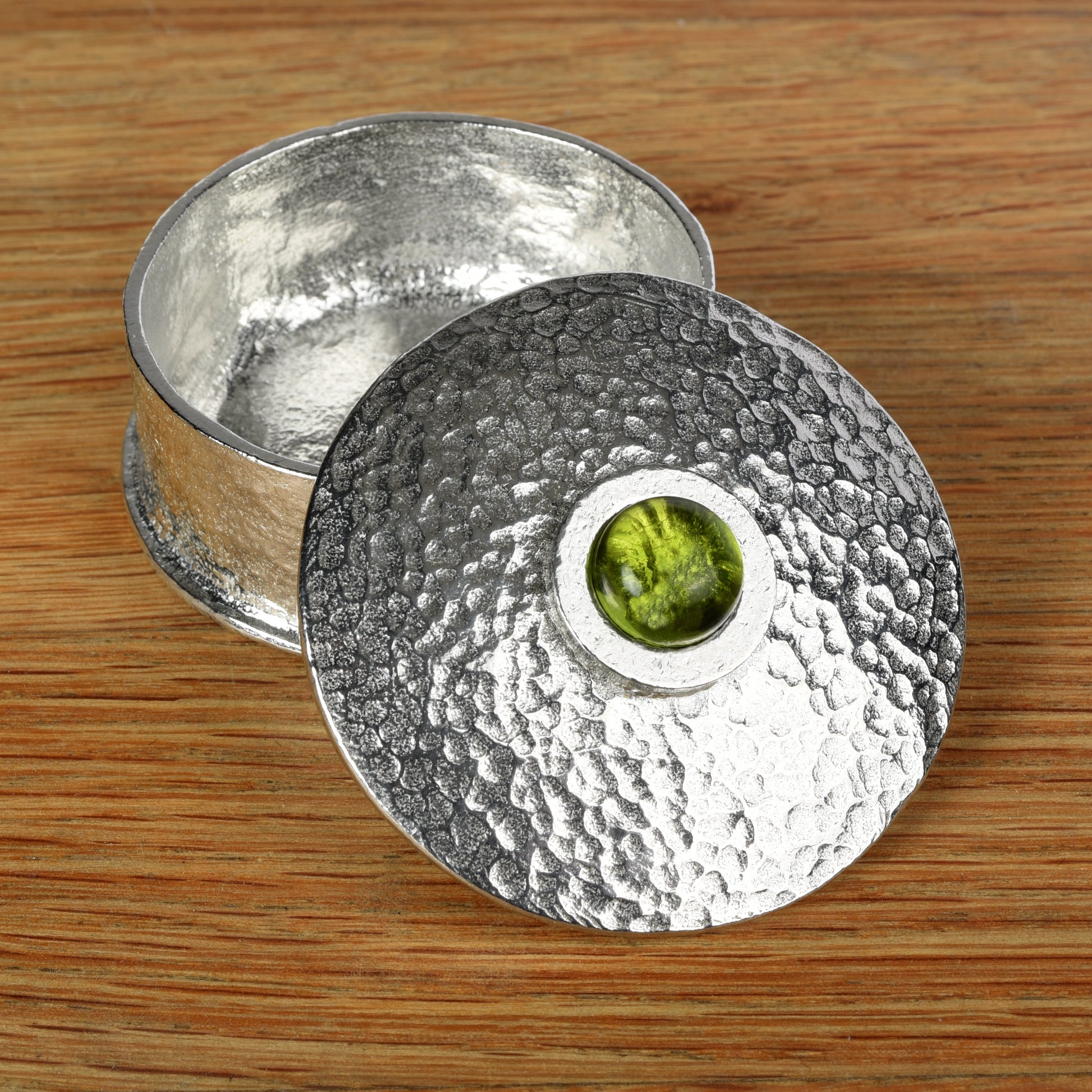 A hammered silver-toned pewter box with its lid leaning against the base, set on a wooden surface. The lid features a circular cutout in the center revealing a peridot stone inlay. August birthstone gifts