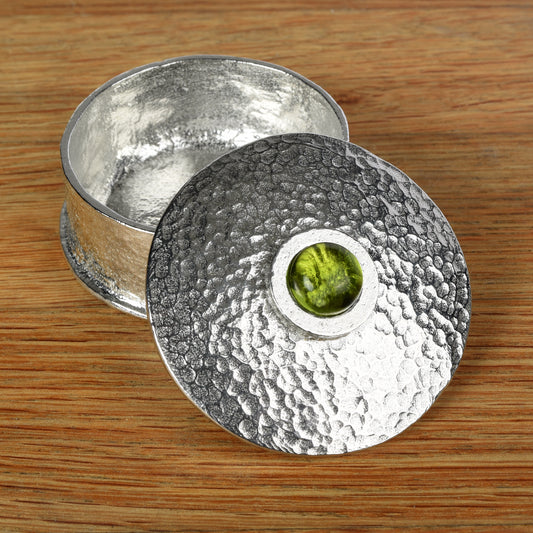 A hammered silver-toned pewter box with its lid leaning against the base, set on a wooden surface. The lid features a circular cutout in the center revealing a peridot stone inlay. August birthstone gifts