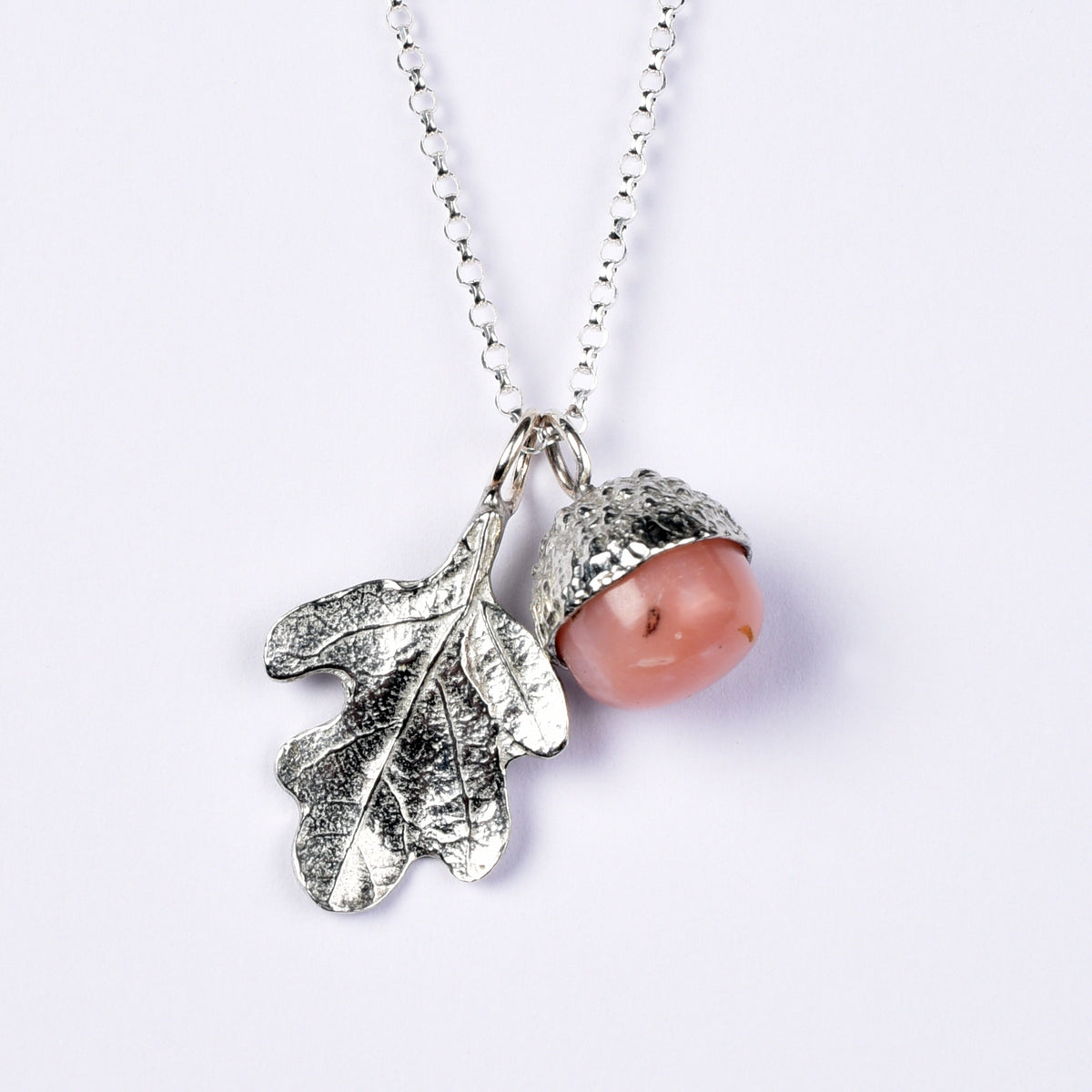 A pewter acorn pendant necklace with a textured silver oak leaf and a smooth, polished pink opal stone bead inside the acorn cap, suspended from a silver belcher chain on a light background.