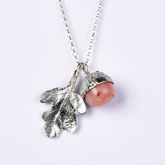 A pewter acorn pendant necklace with a textured silver oak leaf and a smooth, polished pink opal stone bead inside the acorn cap, suspended from a silver belcher chain on a light background.