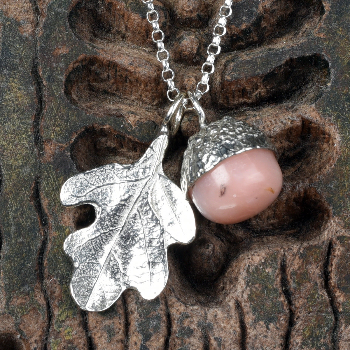 A pewter acorn pendant necklace with a textured silver oak leaf and a smooth, polished pink opal stone bead inside the acorn cap, suspended from a silver belcher chain.