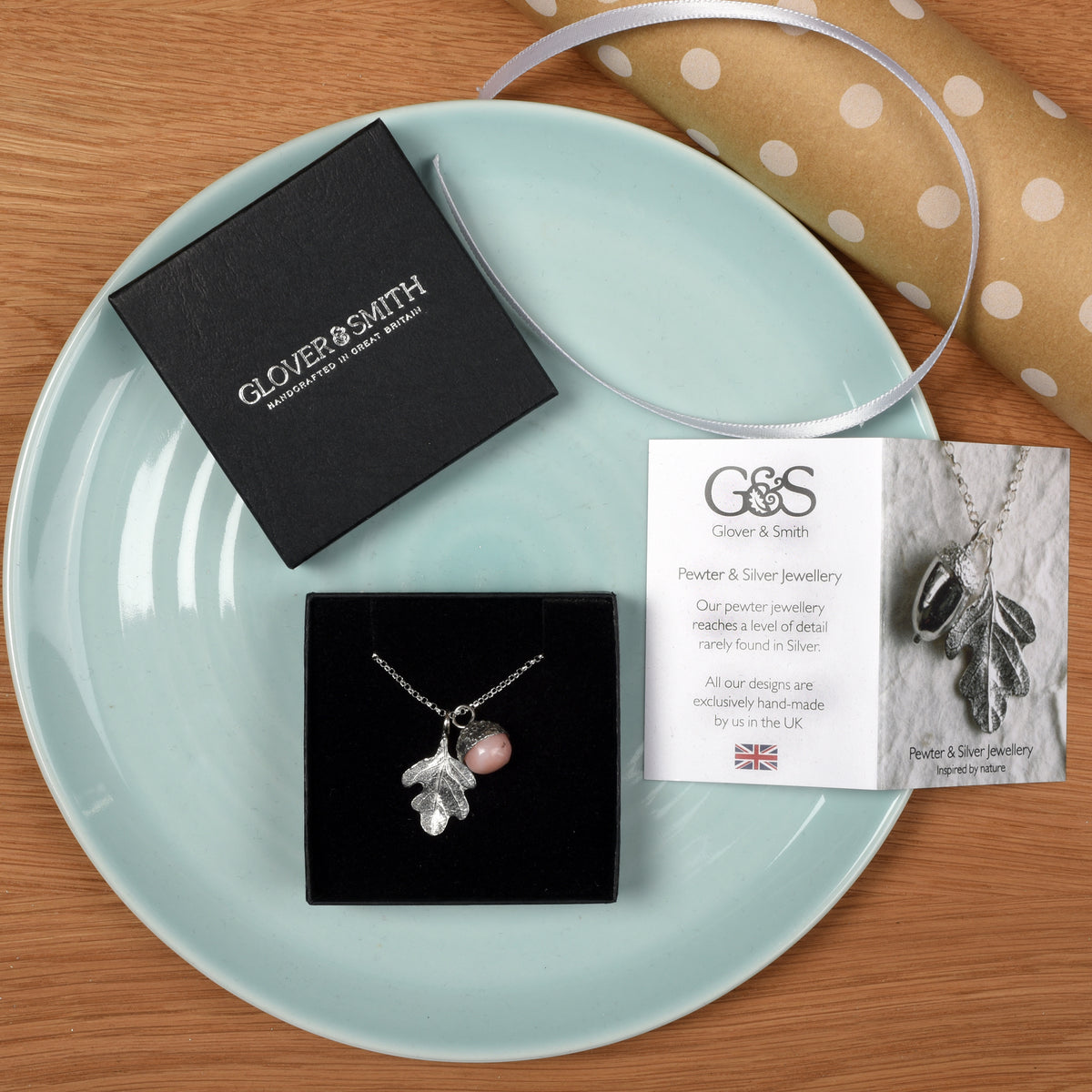 A pewter acorn pendant necklace with a textured silver oak leaf and a smooth, polished pink opal stone bead inside the acorn cap, suspended from a silver belcher chain. Shown in its gift packaging
