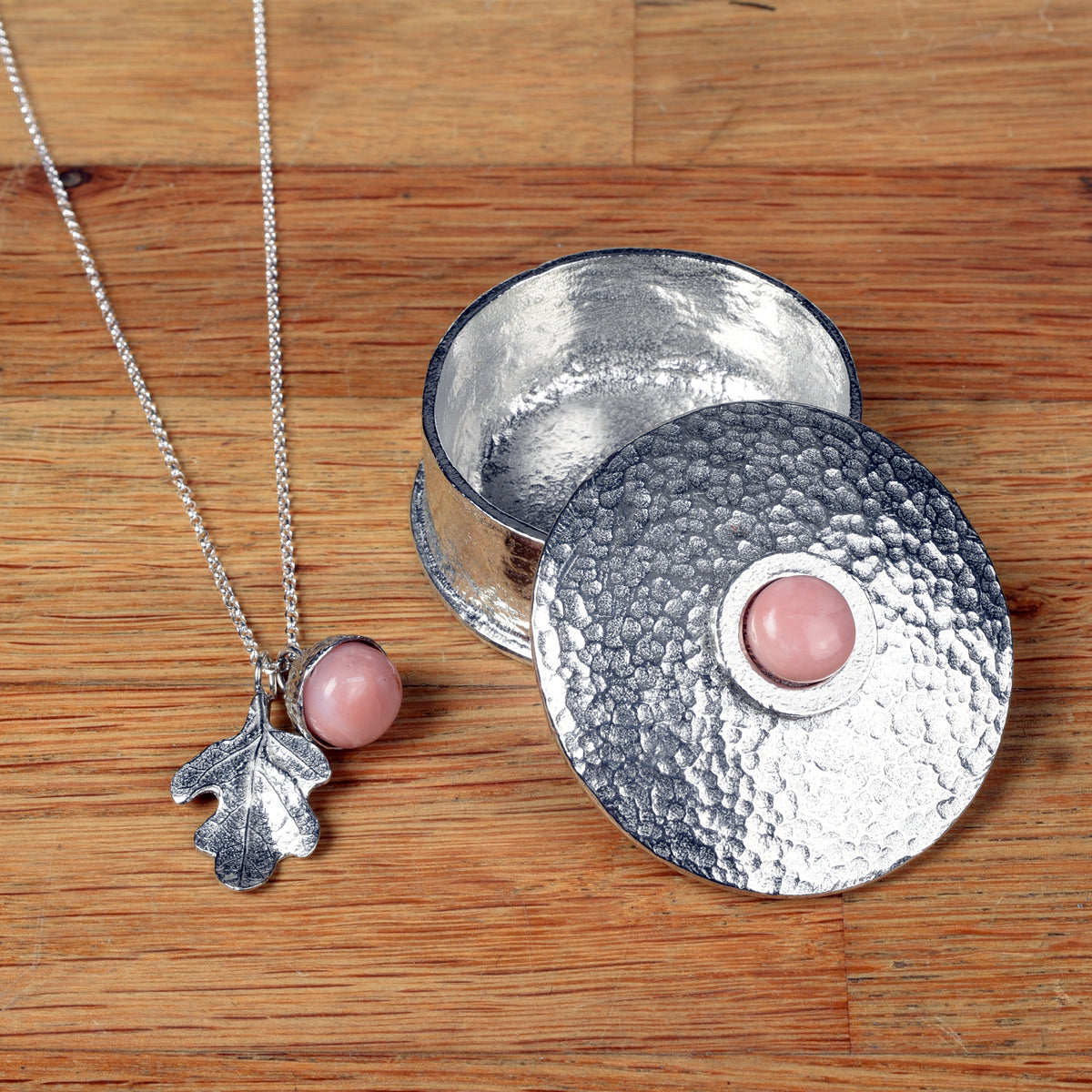 A pewter acorn pendant necklace with a textured pewter oak leaf and a smooth, polished pink opal stone bead inside the acorn cap, shown next to a pewter box with a pink opal on the lid