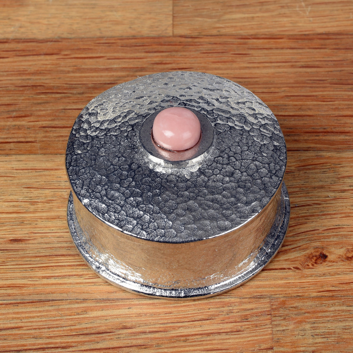 A hammered pewter box with its lid resting on top, featuring a smooth pink opal stone set in the center of the lid, all placed on a wooden surface.