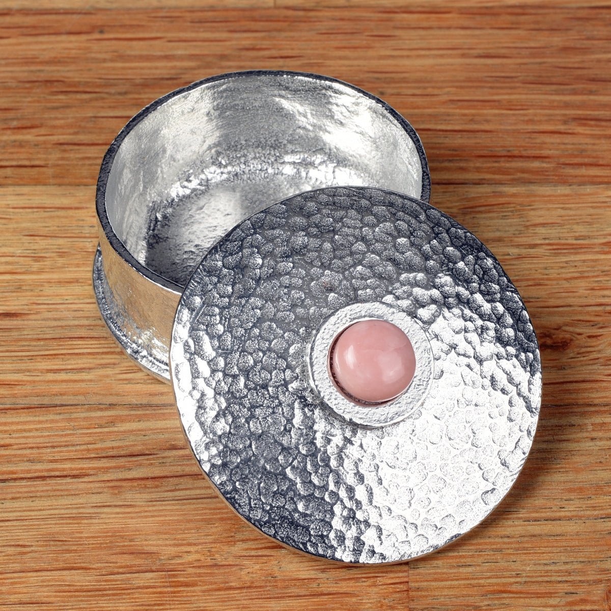 A hammered pewter box with its lid resting on top, featuring a smooth pink opal stone set in the center of the lid, all placed on a wooden surface.