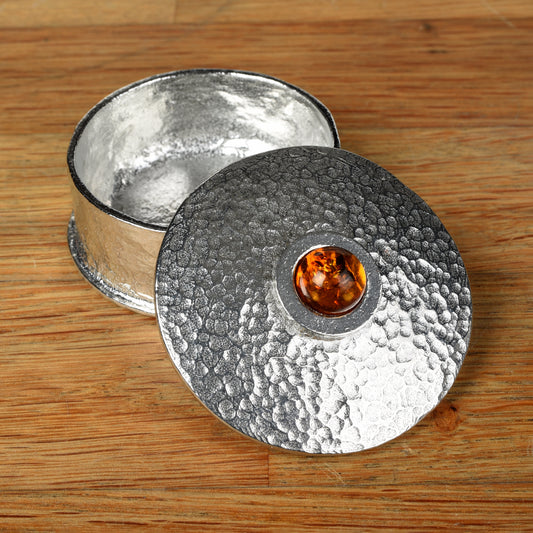 A hammered pewter box with its lid resting on top, featuring a smooth amber set in the center of the lid, all placed on a wooden surface.
