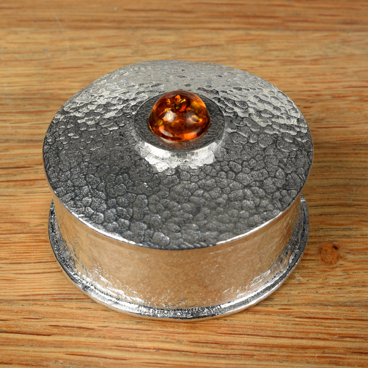 A hammered pewter box featuring a smooth amber set in the center of the lid, all placed on a wooden surface.