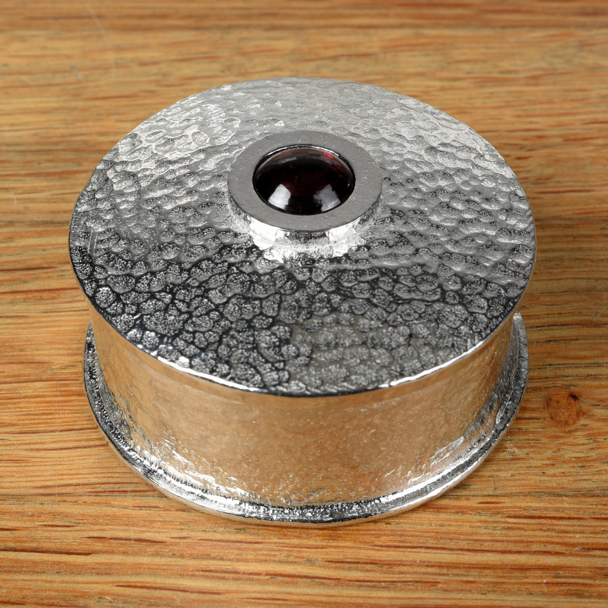 A hammered pewter box featuring a smooth garnet stone set in the center of the lid, all placed on a wooden surface.