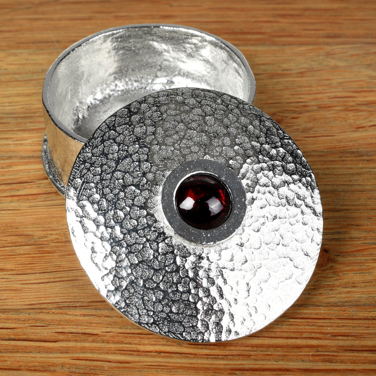 A hammered pewter box with its lid resting on top, featuring a smooth garnet stone set in the center of the lid, all placed on a wooden surface.