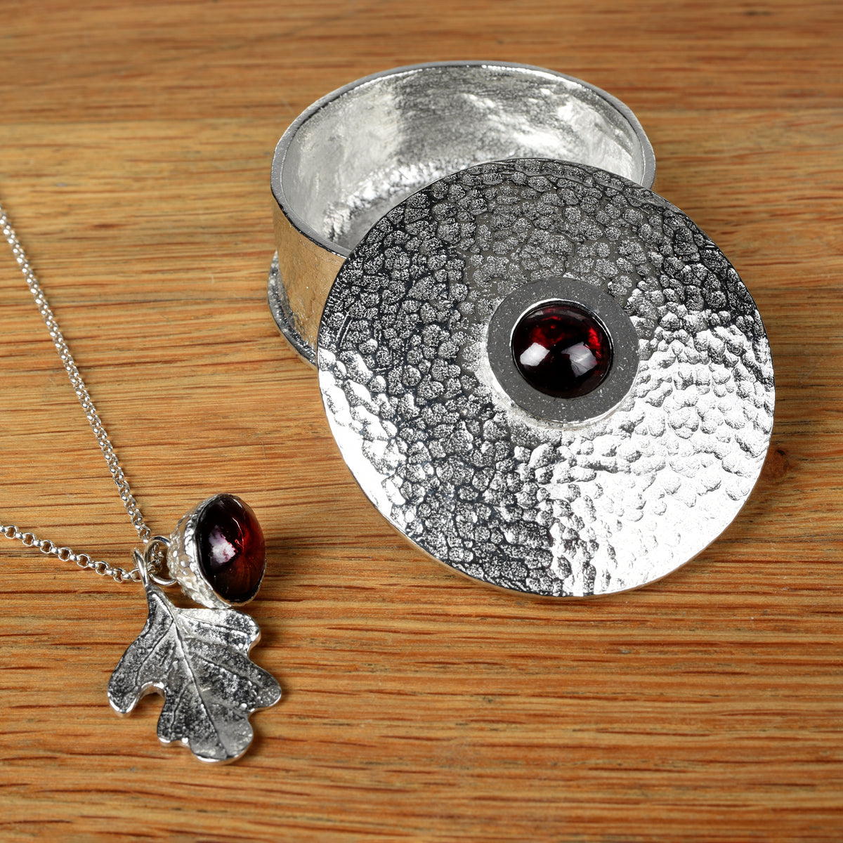 A hammered pewter box with its lid resting on top, featuring a smooth garnet stone set in the center of the lid. Shown with a garnet acorn and oak leaf necklace all placed on a wooden surface.