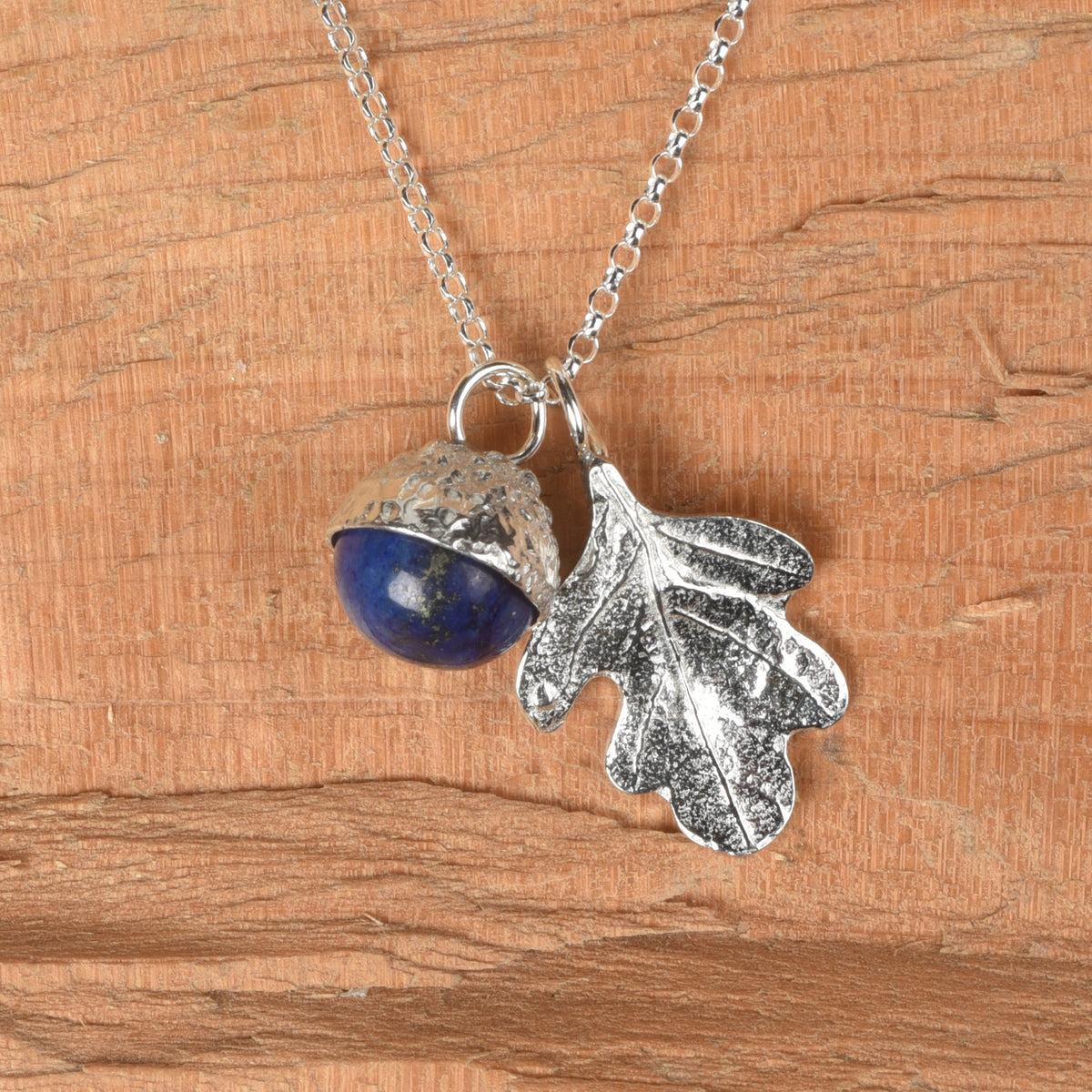 A pewter necklace with a dual pendant featuring a polished blue lapis lazuli stone acorn and a textured oak leaf, displayed on a warm wood grain background.