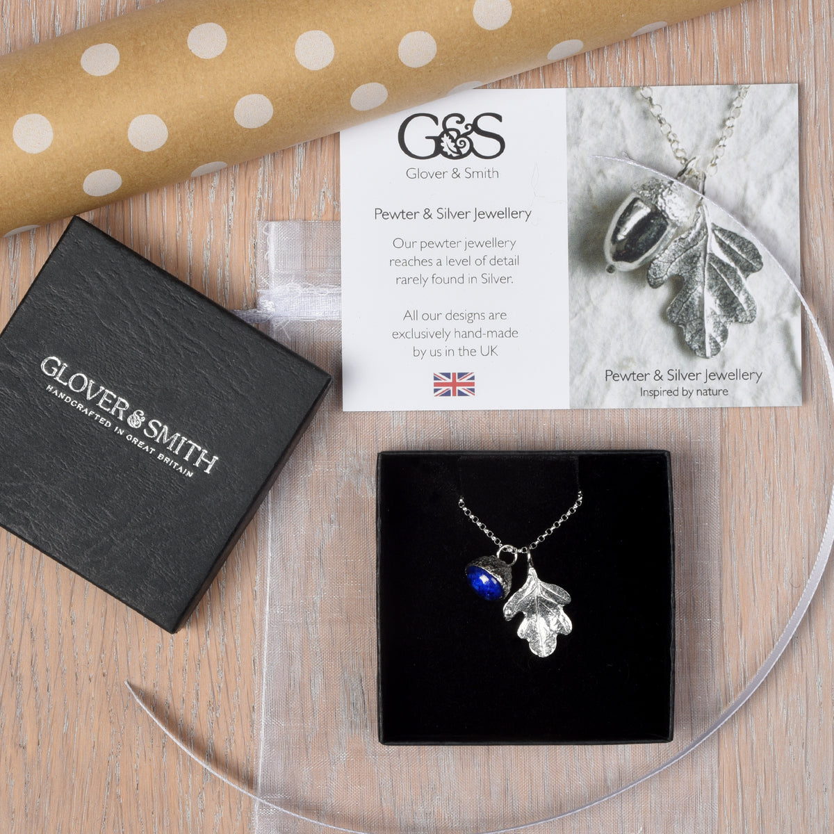 A pewter necklace with an oak leaf and a lapis lazuli acorn shown with its gift packaging, made by Glover and Smith