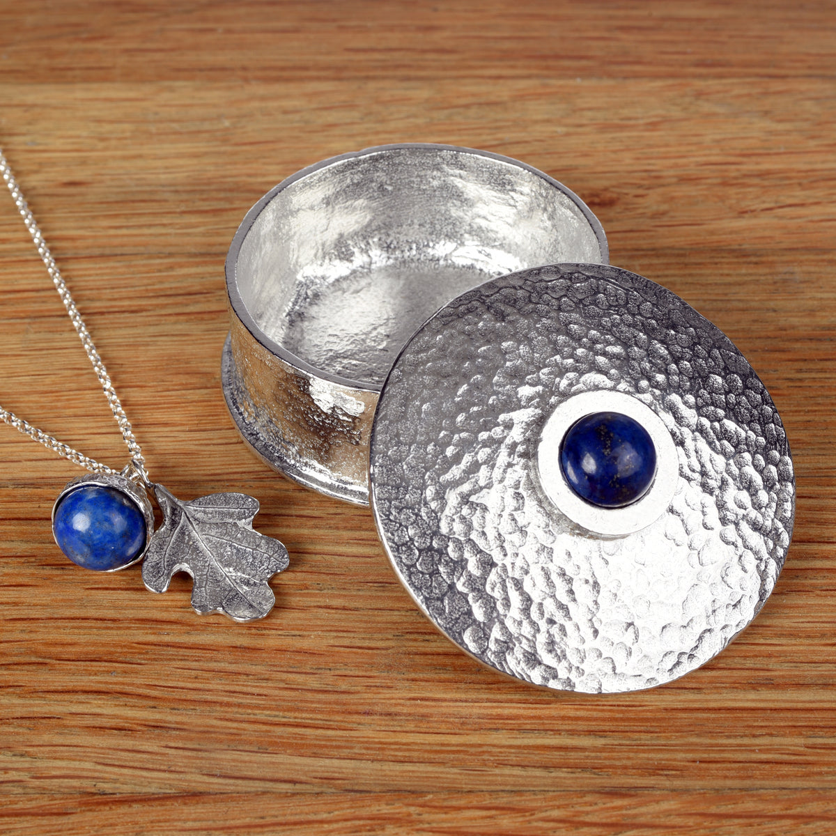 A round pewter box with a lapis lazuli deep blue stone in the centre of the lid, made by Glover and Smith. Next to it a necklace with an oak leaf and a lapis lazuli acorn 