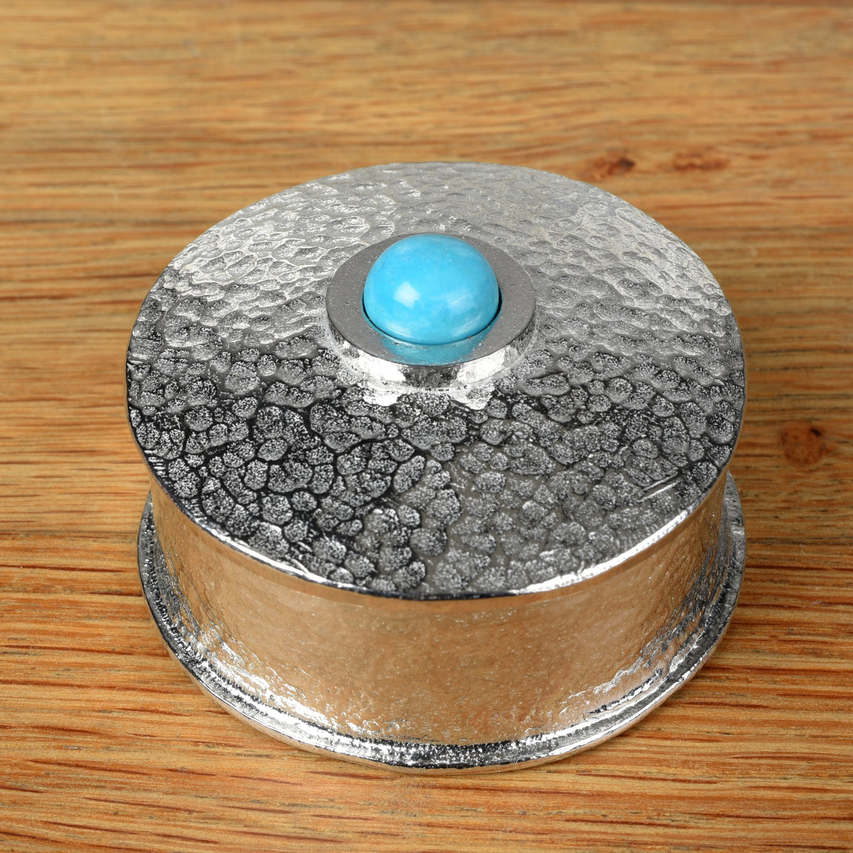 A hammered pewter box with its lid featuring a smooth turquoise set in the center of the lid, all placed on a wooden surface.