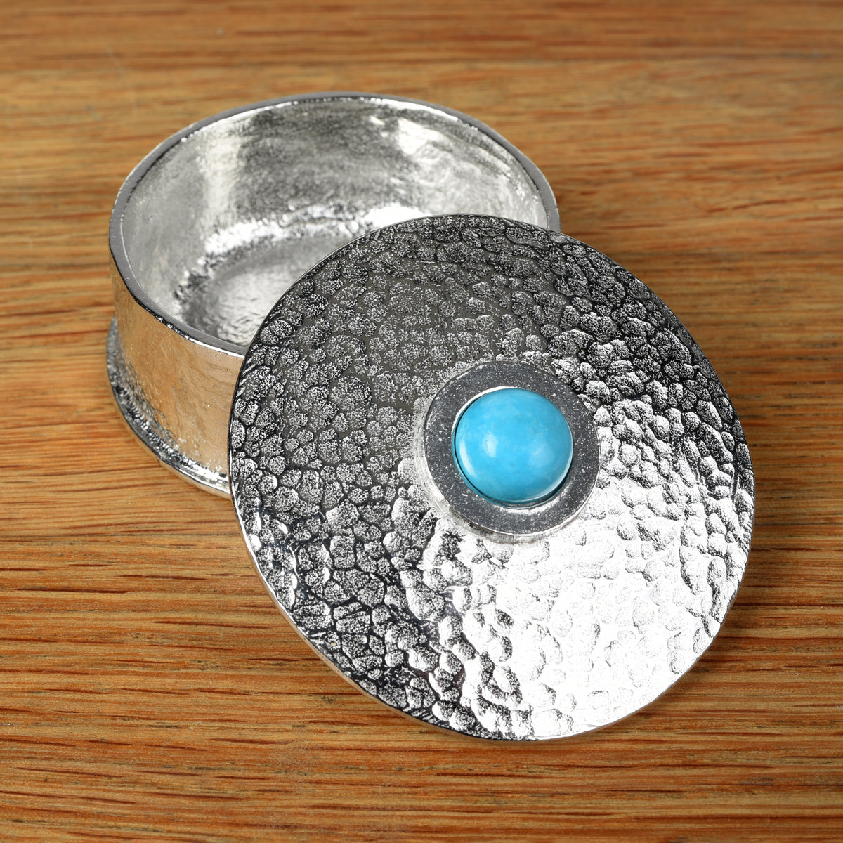 A hammered pewter box with its lid resting on top, featuring a smooth turquoise set in the center of the lid, all placed on a wooden surface.