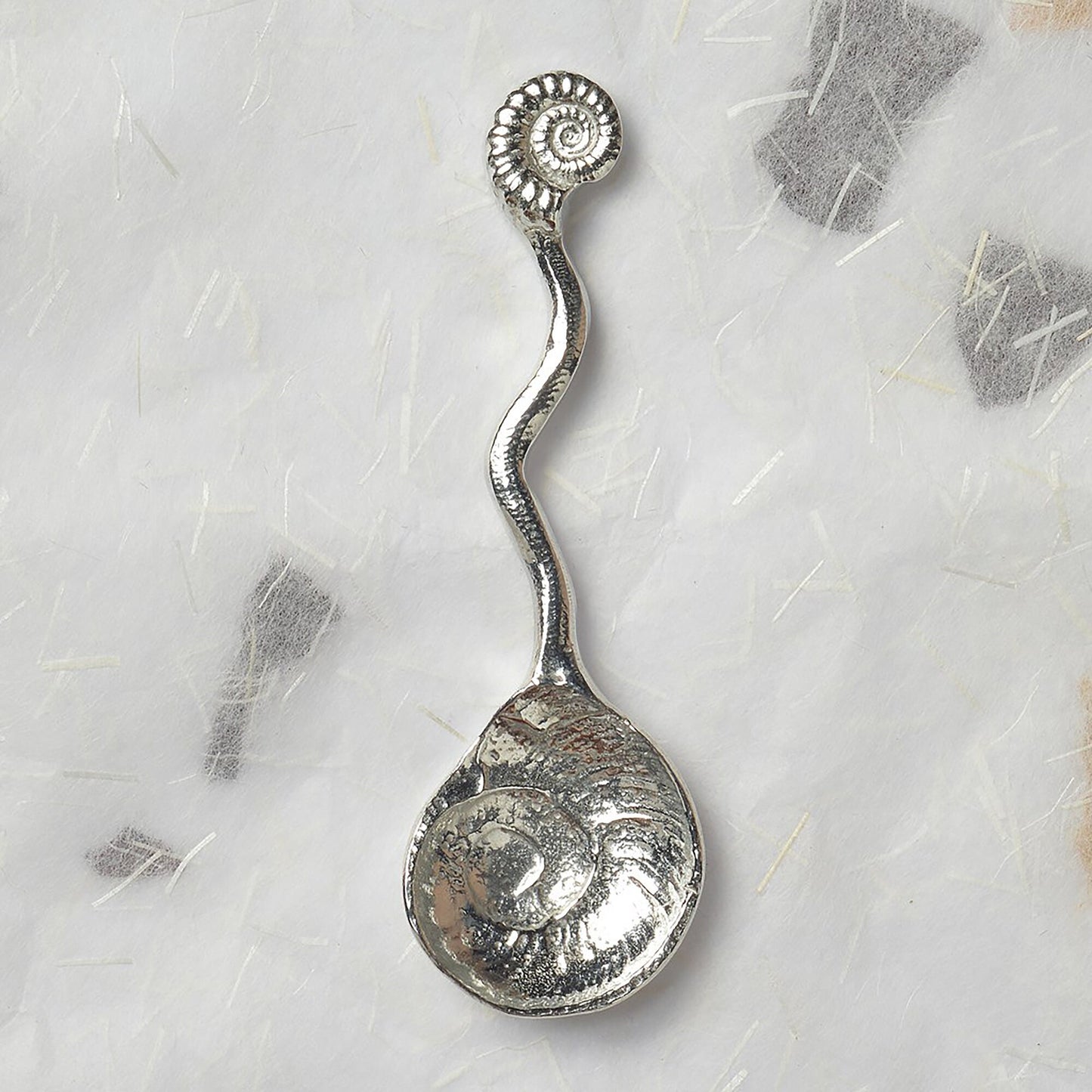 A small spoon pewter sugar spoon. A shell shaped bowl leads around a wavy stem to a perfectly formed ammonite fossil. Practical & useful geology gifts for fossil lovers. UK handmade