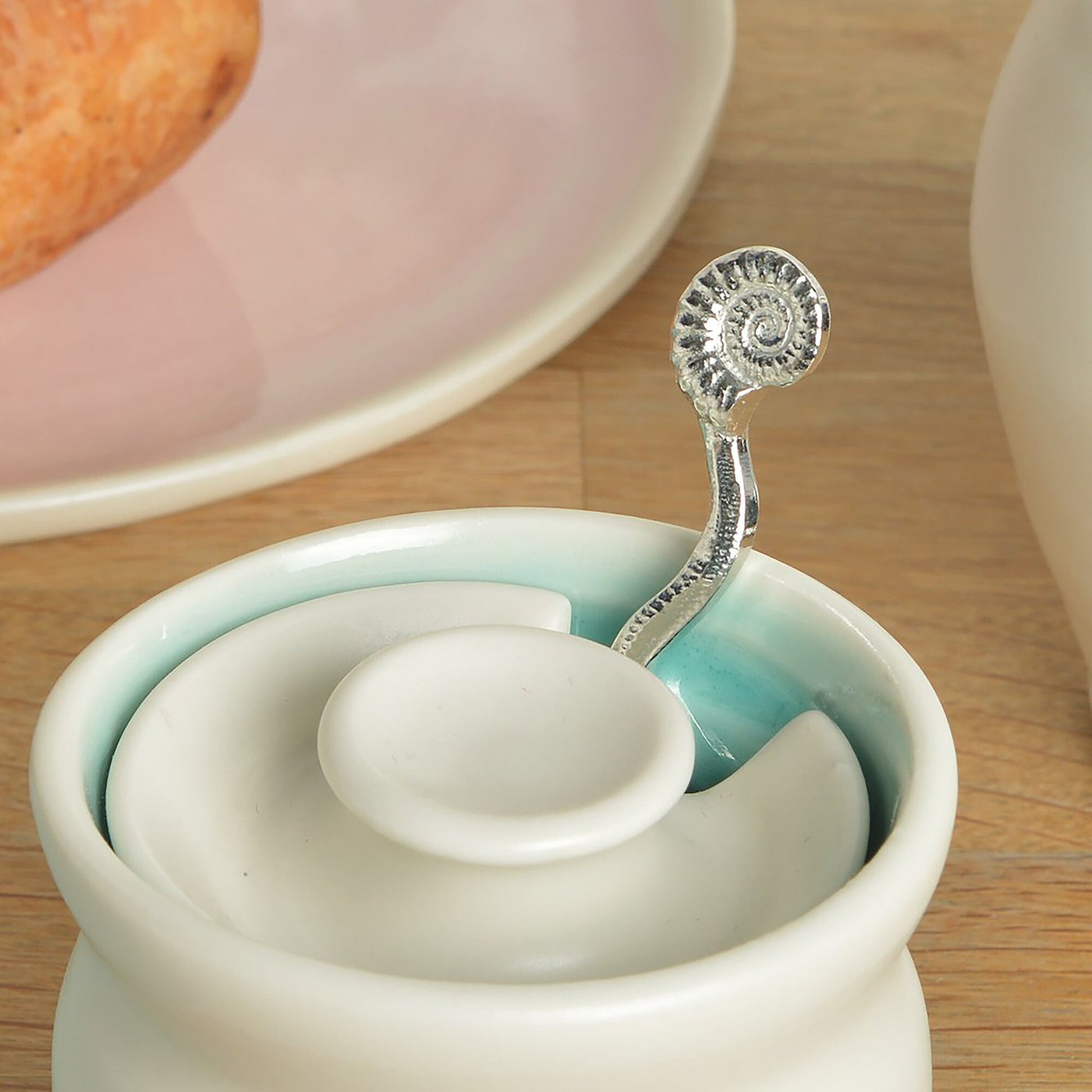 A small spoon pewter sugar spoon. A shell shaped bowl leads around a wavy stem to a perfectly formed ammonite fossil. Practical & useful geology gifts for fossil lovers. UK handmade