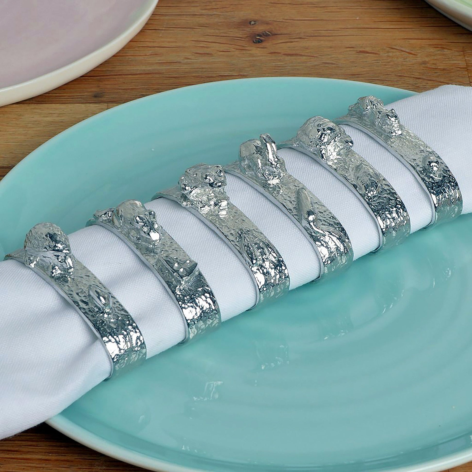 A set of 6 textured pewter oval-shaped napkin rings. Each one has a sculpture of an animal persuing its favourite food. A hedgehog and snail, a mouse and cheese, a frog and insect, a rabbit and carrot, a tortoise and leaf and a squirrel and acorn.