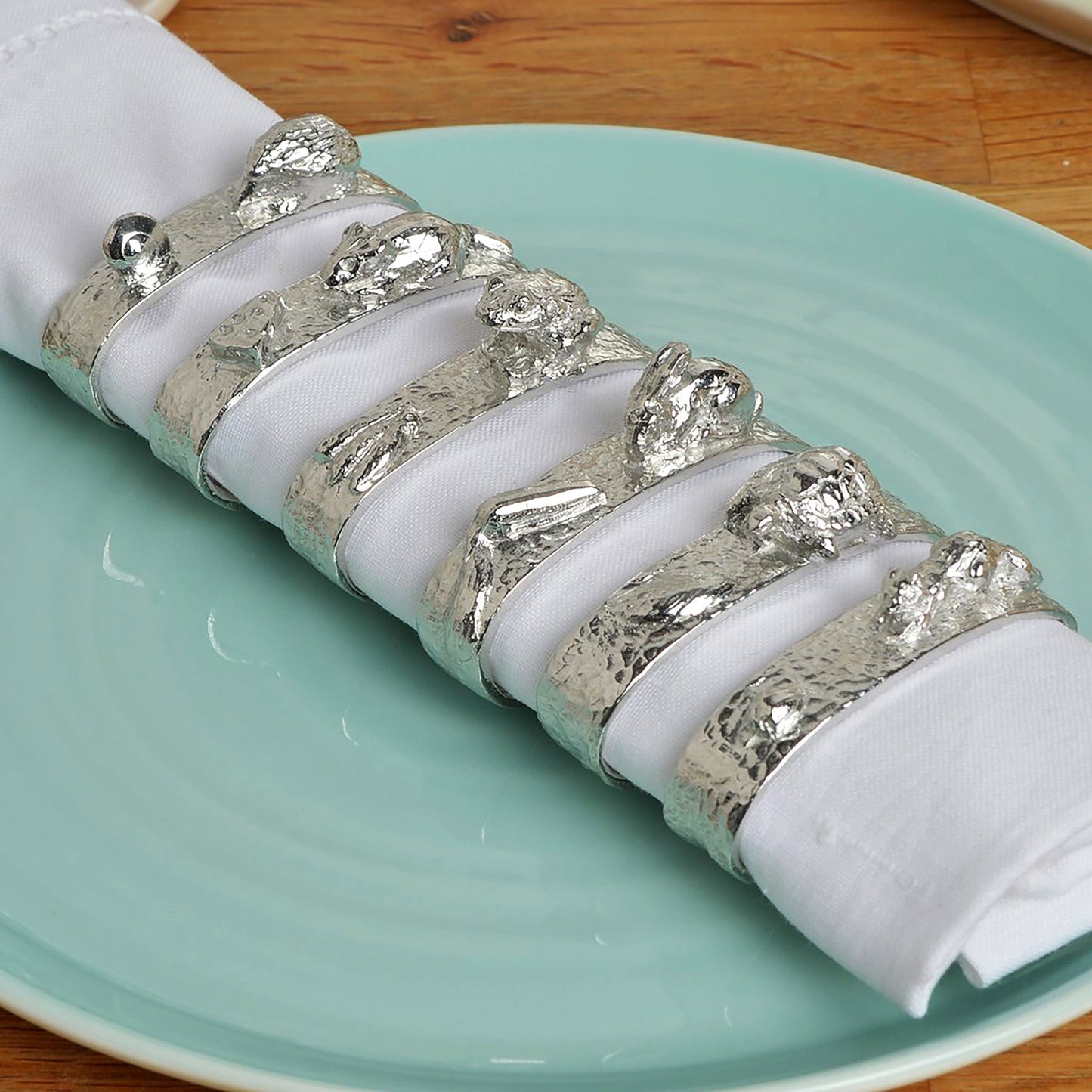 A set of 6 textured pewter oval-shaped napkin rings. Each one has a sculpture of an animal persuing its favourite food. A hedgehog and snail, a mouse and cheese, a frog and insect, a rabbit and carrot, a tortoise and leaf and a squirrel and acorn.